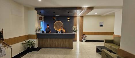 A hotel reception area with a reception desk, two receptionists, a waiting area with chairs, and a potted plant.