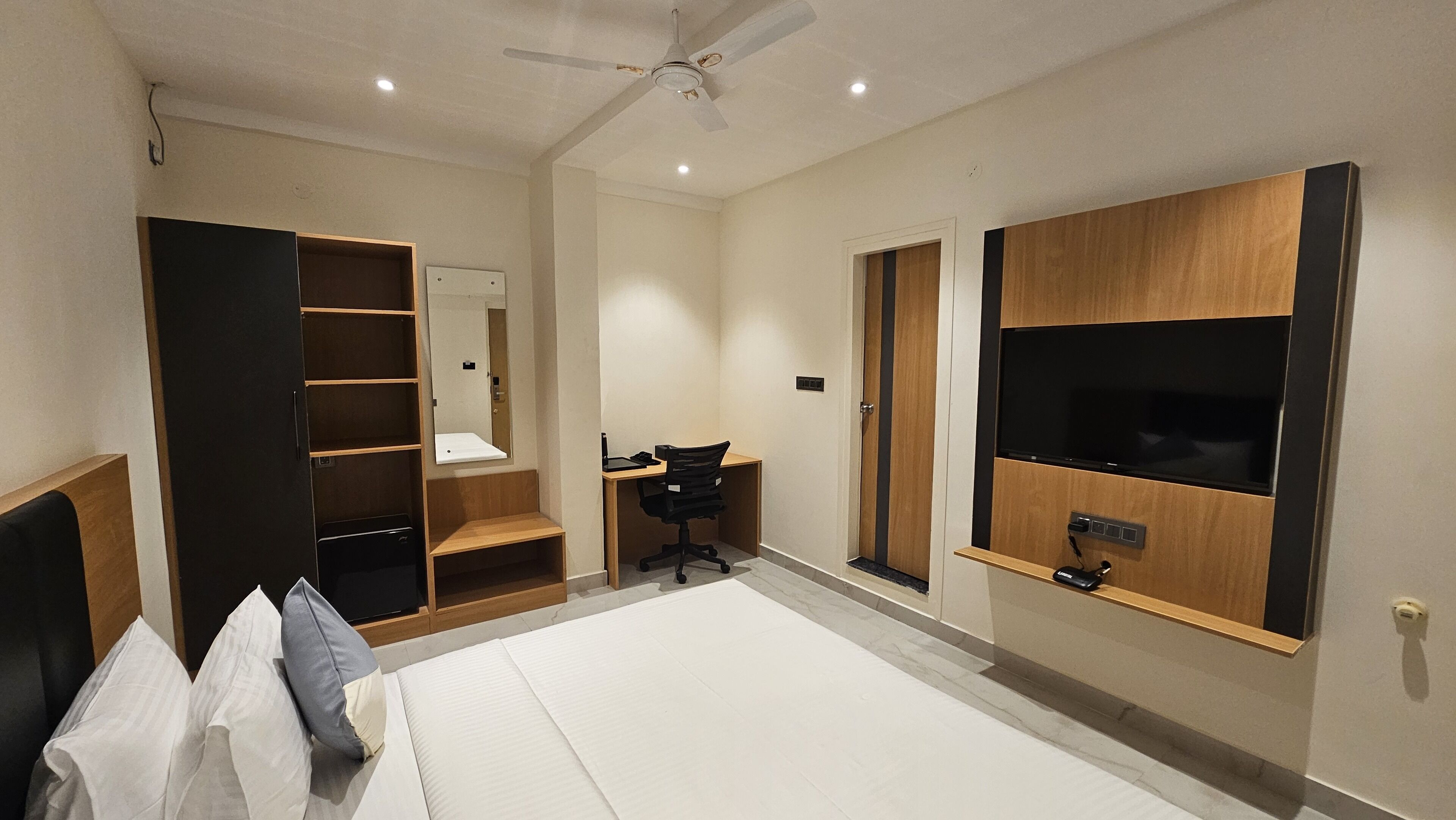 A bedroom with a bed, a desk, a chair, a wardrobe, a TV mounted on the wall, and a ceiling fan.