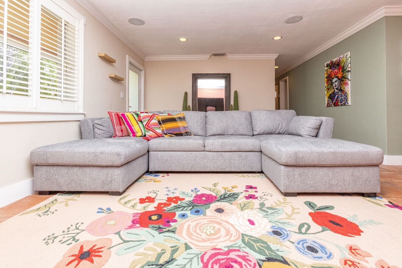 Un sofá seccional gris con almohadones de colores, un tapete estampado con motivos florales y una puerta que da a un jardín al fondo.