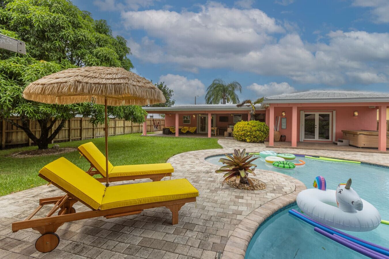 Ein Garten mit Pool, gelben Liegestühlen, einem Strohdachschirm und einem Haus mit rosa Fassade.