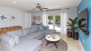 A living room with a grey sectional sofa, a round coffee table, a ceiling fan, and a pool visible through the glass door.