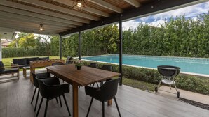 An outdoor patio with a wooden table and chairs, a foosball table, and a barbecue grill, overlooking a swimming pool.