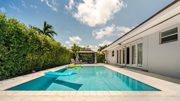 A swimming pool with a tiled deck, surrounded by a hedge and a building with large windows. - Hawaii Paradise on Miami 4BR Big Pool (Miami)