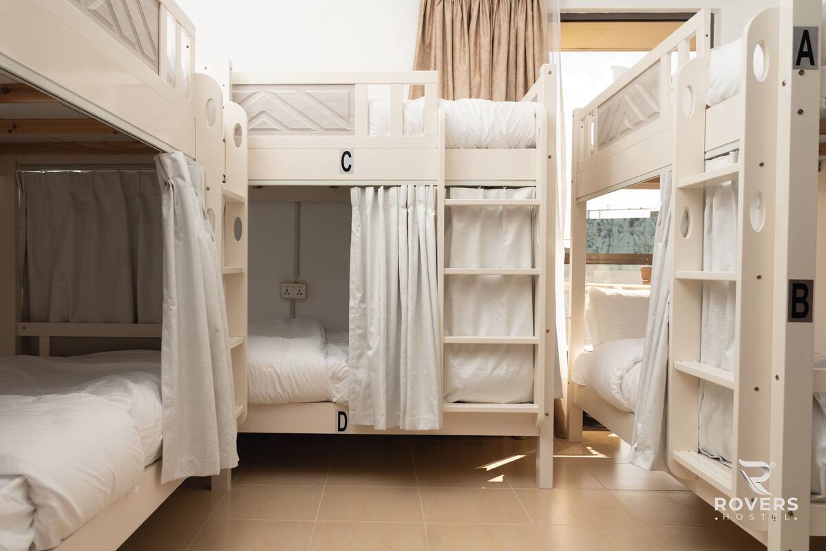 Une chambre de dortoir équipée de lits superposés étiquetés A, B, C et D, d’un petit placard et d’une fenêtre avec des rideaux.