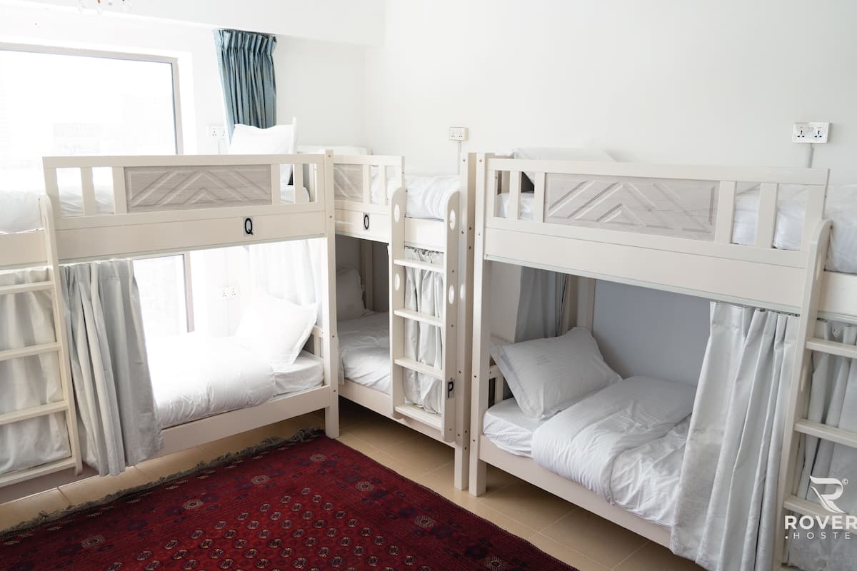 Une chambre avec un lit superposé, un tapis rouge à motifs, du linge de lit blanc et une fenêtre avec des rideaux.
