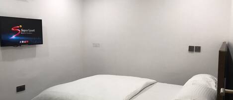 A single bed with white bedding in a room with white walls, a mounted TV displaying the SuperSport logo, and a ceiling light.