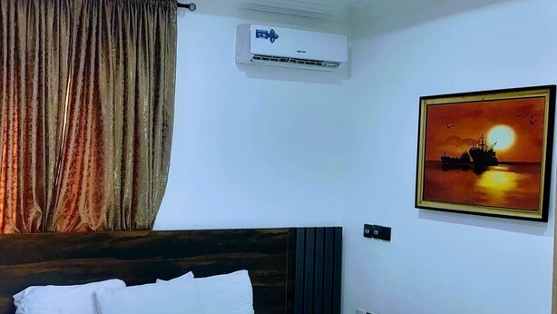 A hotel room with a large bed, an air conditioning unit, a painting of a sunset, and a curtain. -  Skenyo Hotel & Apartments (Abuja)