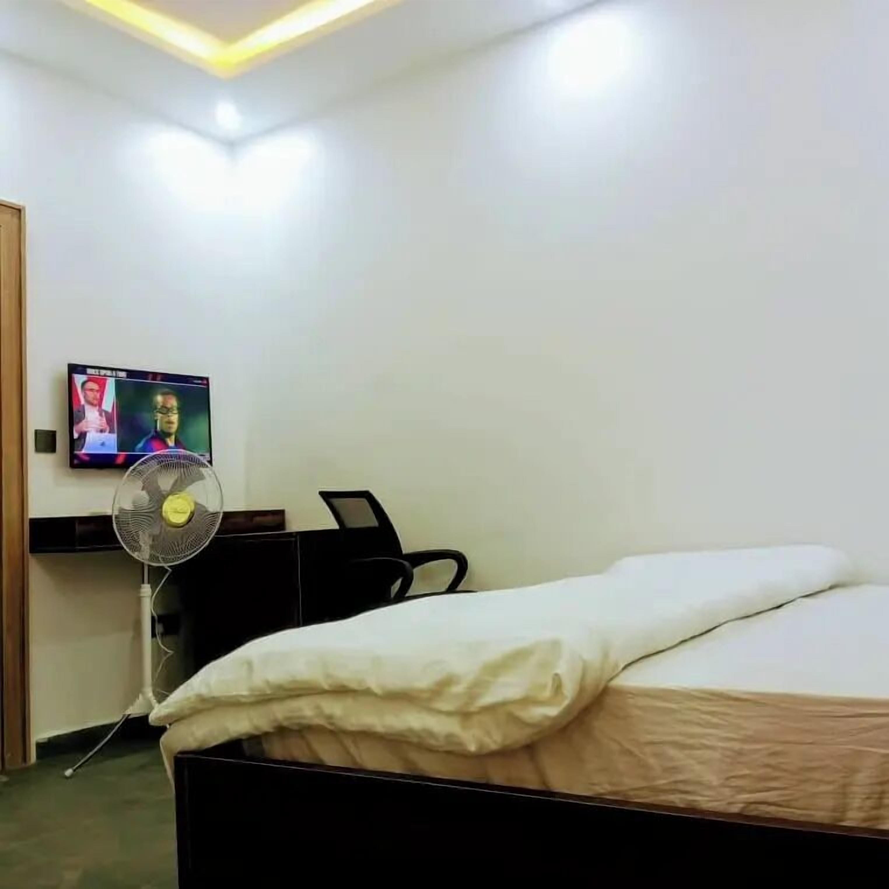 A hotel room with a bed, a desk with a computer, a chair, and a TV displaying a show.