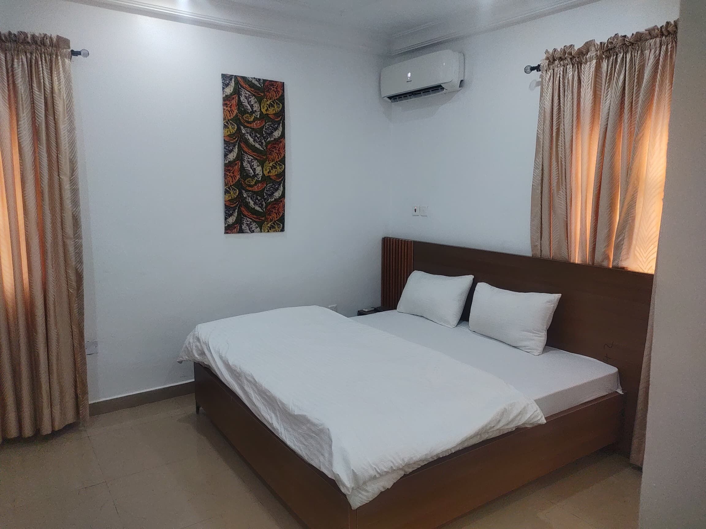 A bedroom with a single bed, beige curtains, a wall-mounted air conditioner, and a colorful abstract painting.