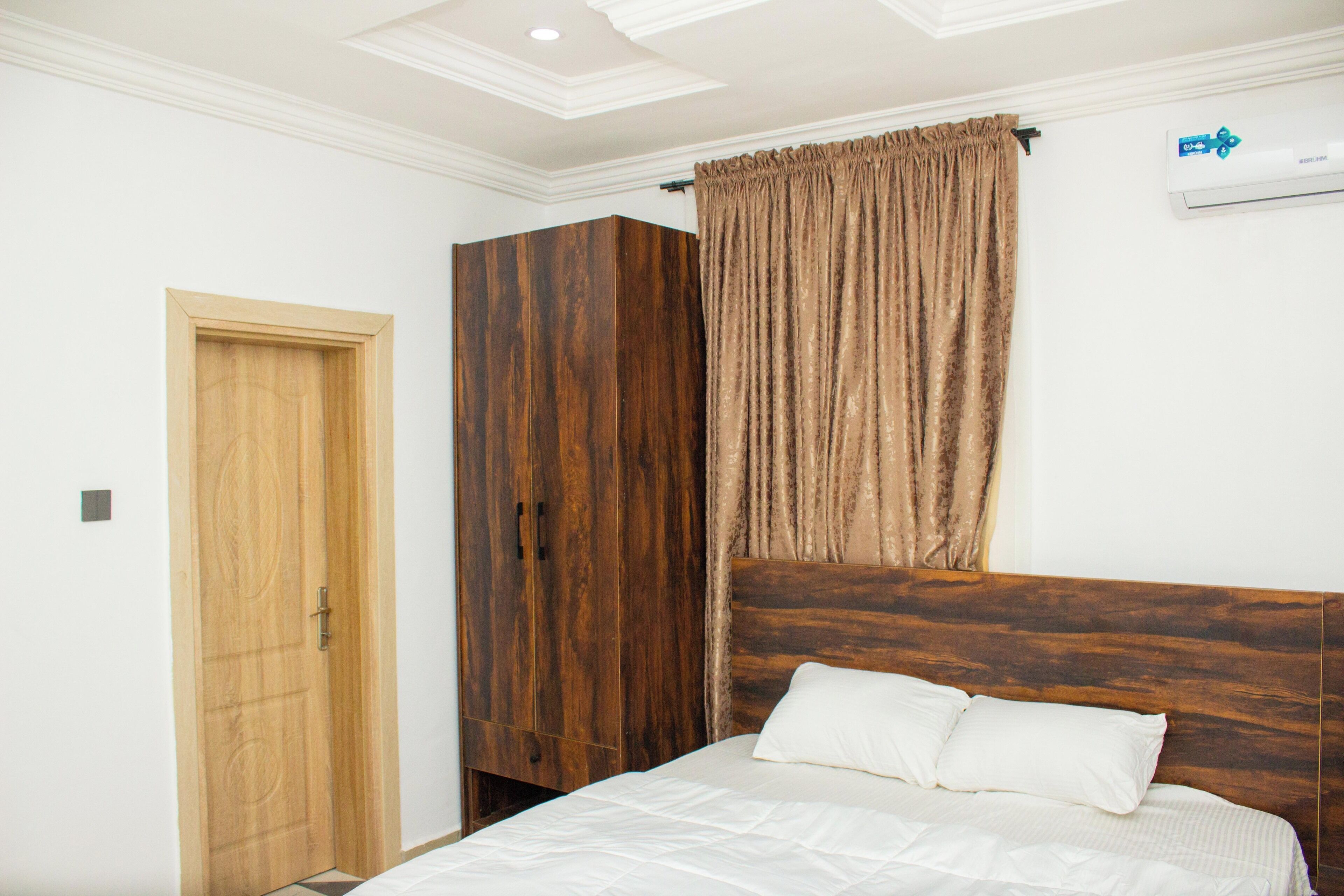 A bedroom with a wooden headboard, a wooden wardrobe, a white bed with two pillows, a wooden door, and a window with curtains.