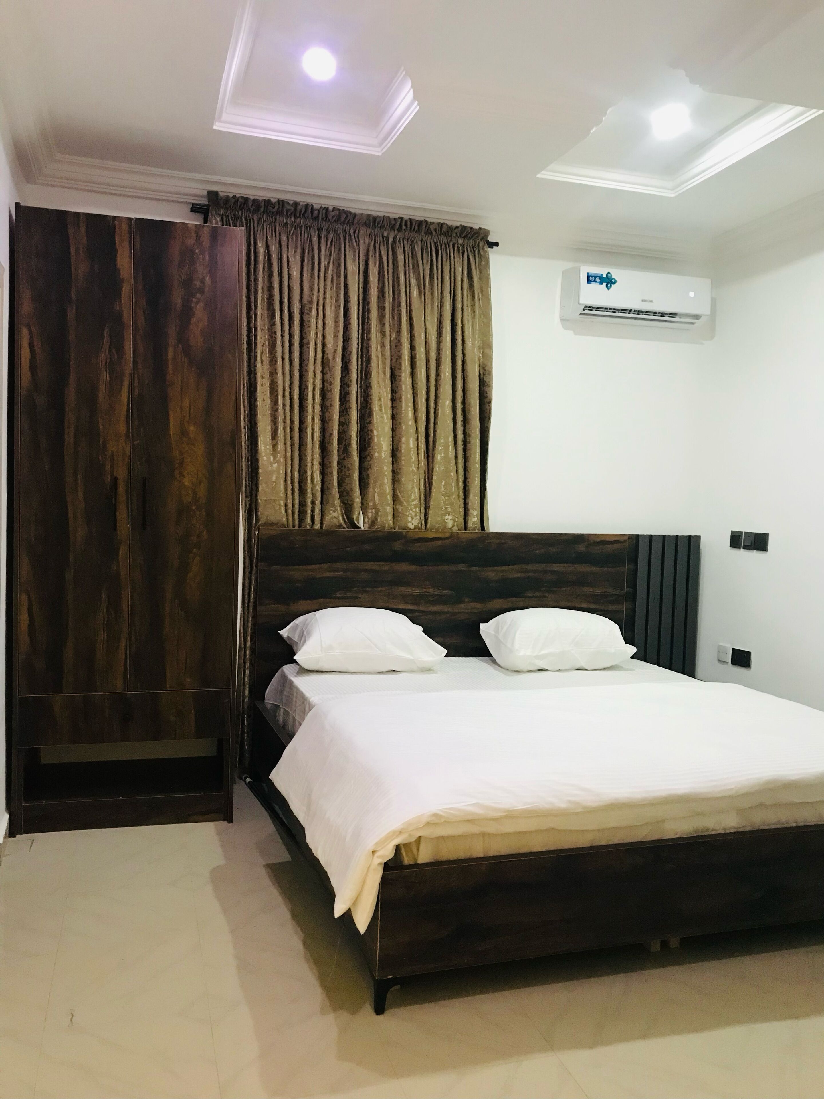 A bedroom with a dark wooden bed, white bedding, a large window with curtains, and an air conditioning unit on the wall.