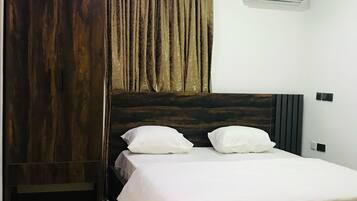 A bedroom with a dark wooden bed, white bedding, a large window with curtains, and an air conditioning unit on the wall.
