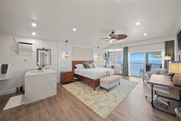 A modern hotel room with a wooden floor, a large bed with a headboard, a desk with a chair, a balcony with a view, and a ceiling fan. - Hamakua Hotel (Honomu)
