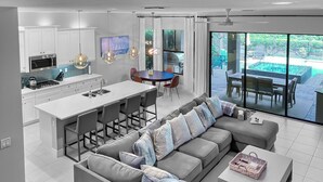 A modern kitchen with white cabinets, a central island, and pendant lighting. There is a dining area with a round table and chairs, and a large window offering a view of the outdoors.