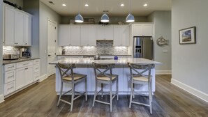 Une cuisine moderne avec des meubles blancs, un grand îlot central doté de tabourets, un réfrigérateur en acier inoxydable et un éclairage suspendu.
