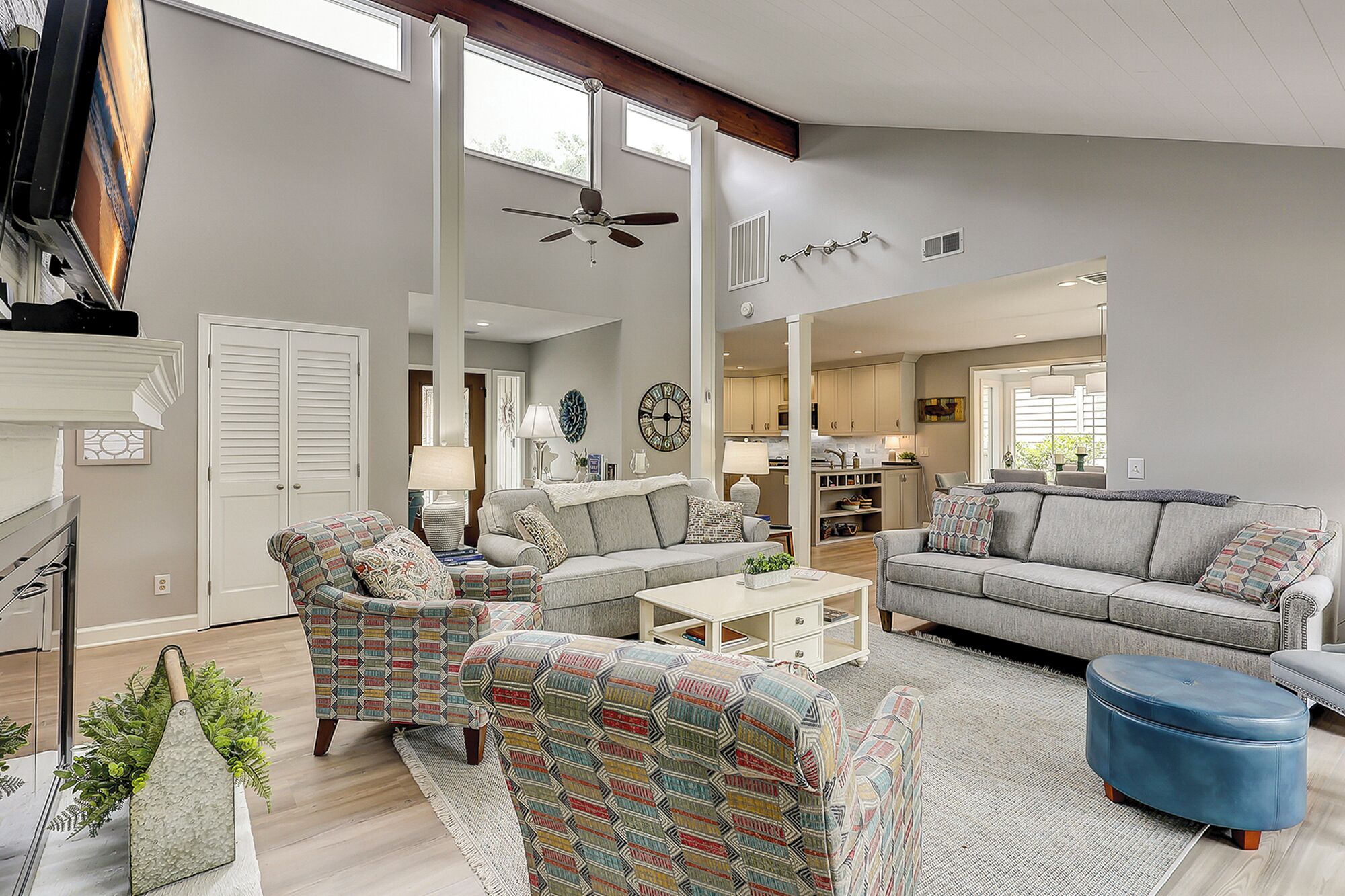 A spacious living room with a grey sofa, patterned armchairs, a coffee table, and a blue ottoman. A ceiling fan and a large window provide natural light.