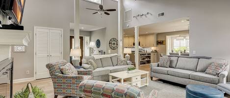 A spacious living room with a grey sofa, patterned armchairs, a coffee table, and a blue ottoman. A ceiling fan and a large window provide natural light.