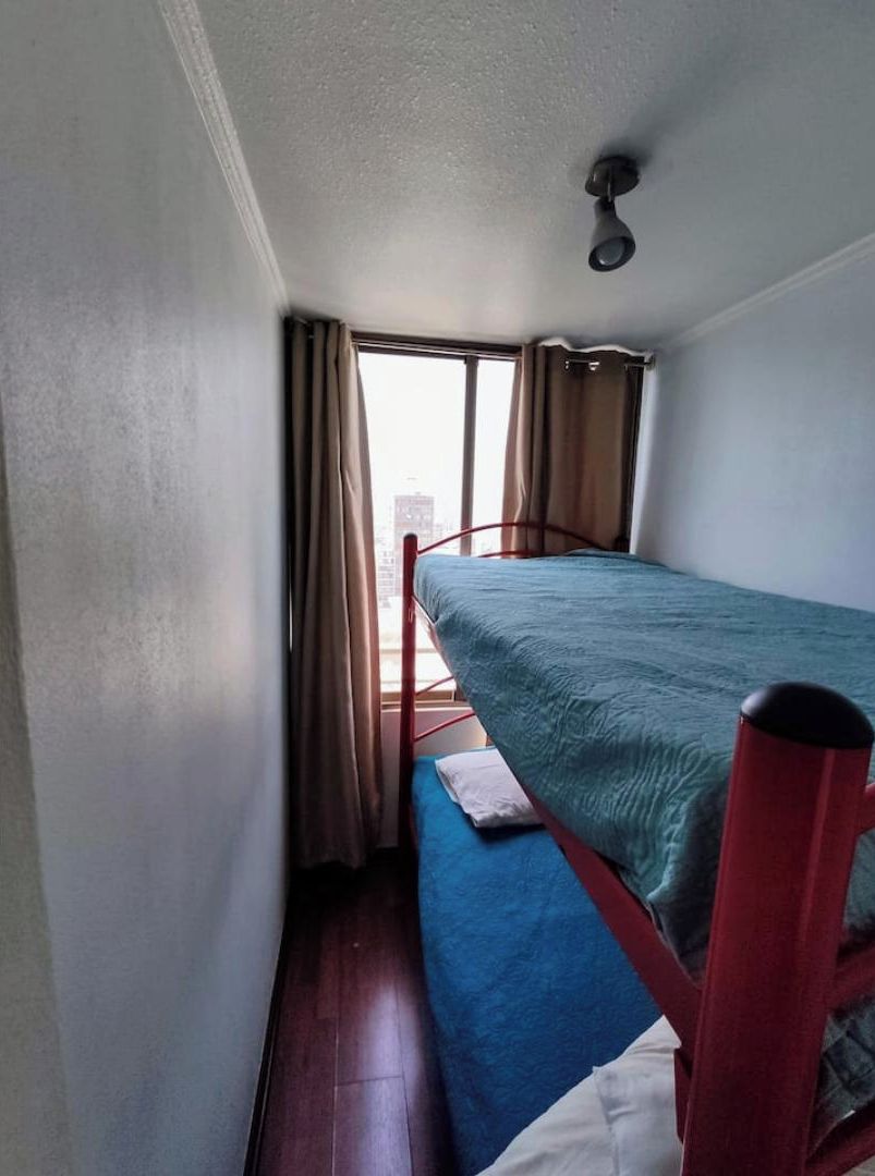 A small, single-bed room with a window, a red metal bed frame, and a white wall.