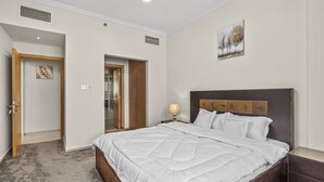 A bedroom with a large bed, wooden headboard, bedside table with lamp, and a painting on the wall. - Pure Living - Cozy Apartment With Balcony In Silicon Oasis (Dubai)