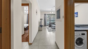 A hallway leading to a living area with a washing machine, a sofa, and a TV. - Pure Living - Cozy Apartment With Balcony In Silicon Oasis (Dubai)