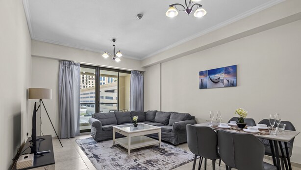 A modern living room with a grey sofa, a coffee table, and a dining area with chairs and a table set for a meal. - Pure Living - Cozy Apartment With Balcony In Silicon Oasis (Dubai)