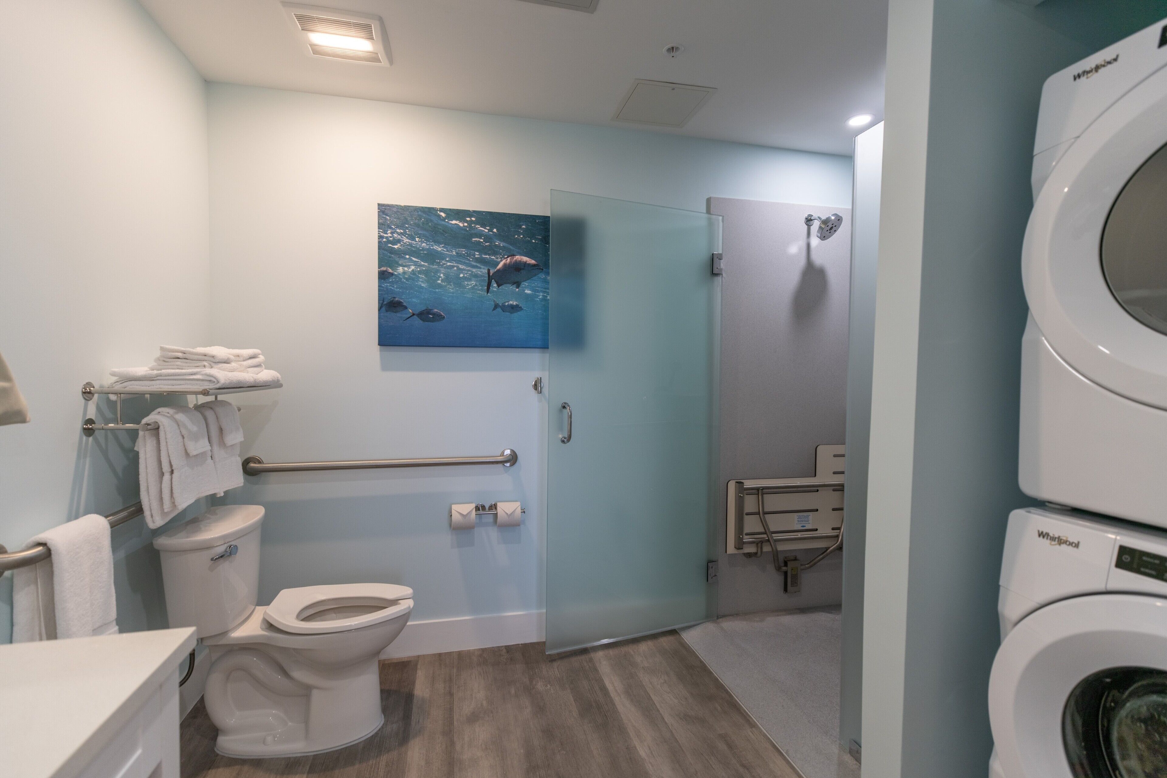 A modern bathroom with a toilet, a glass shower enclosure, a Whirlpool washing machine, and a painting of marine life on the wall.