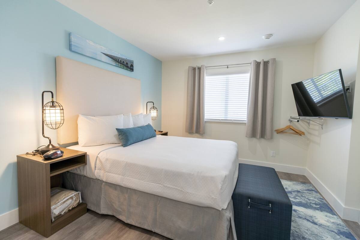 Un dormitorio ordenado con cama, mesitas de noche, televisor y ventana con cortinas.