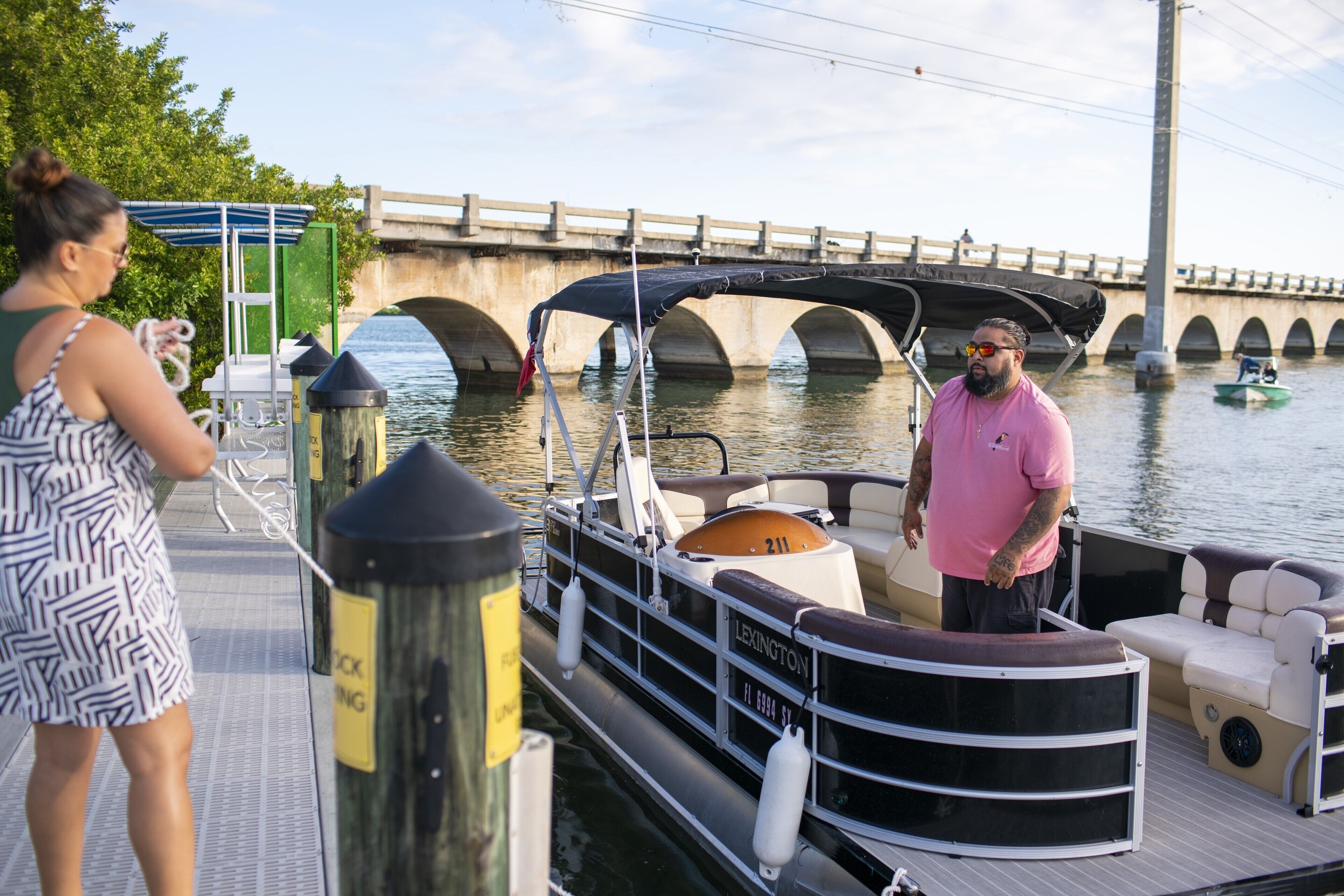 A boat docked at a pier with a covered seating area and a bridge in the background.