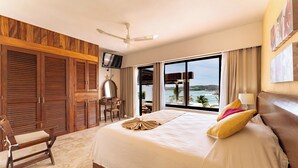 A hotel room with a large bed, a desk, a chair, a ceiling fan, and a view of the ocean.
