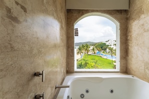 Une salle de bain avec une baignoire, une fenêtre offrant une vue en arc de cercle sur une piscine et la verdure environnante, et un mur en pierre.