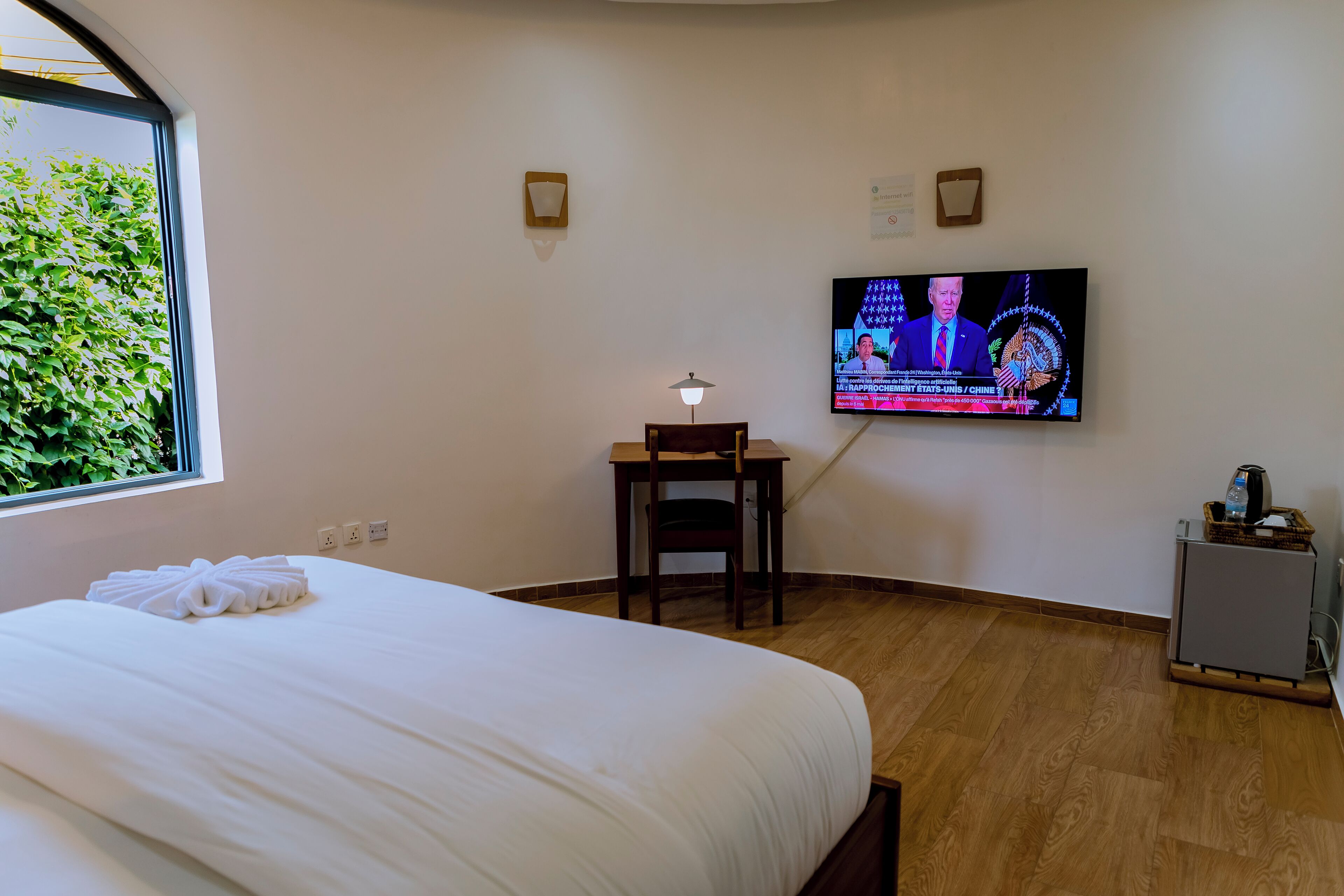 A hotel room with a bed, a desk, a TV mounted on the wall, and a window with a view of greenery.