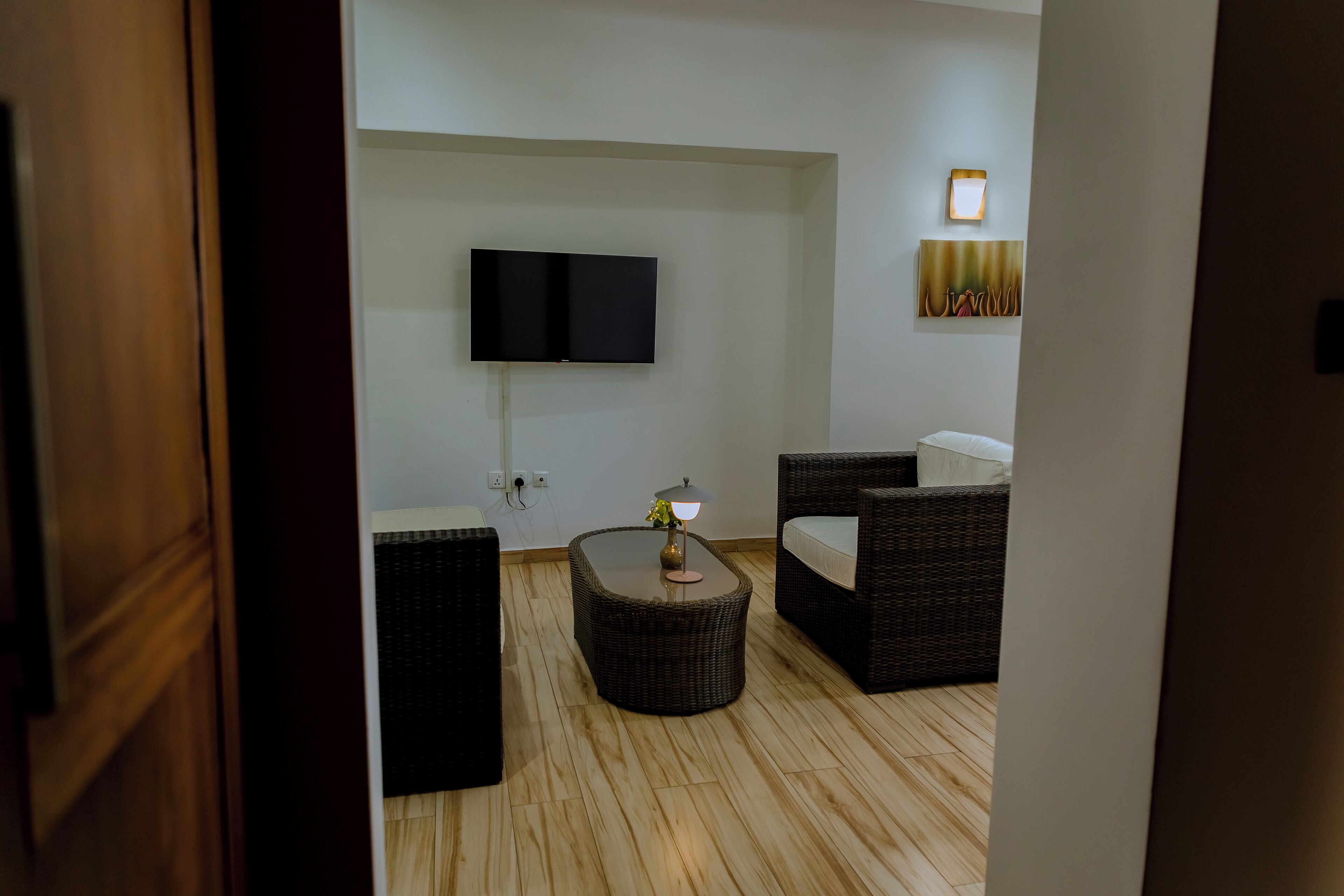 A modern living room with a flat-screen TV mounted on the wall, a round coffee table with a lamp, and two wicker sofas.