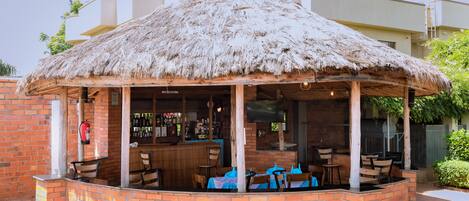 A covered outdoor seating area with brick walls, a thatched roof, and a bar with visible bottles.