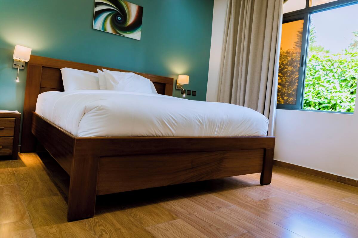 A neatly made bed with white linens in a room with teal walls, a wooden floor, and a window with curtains.