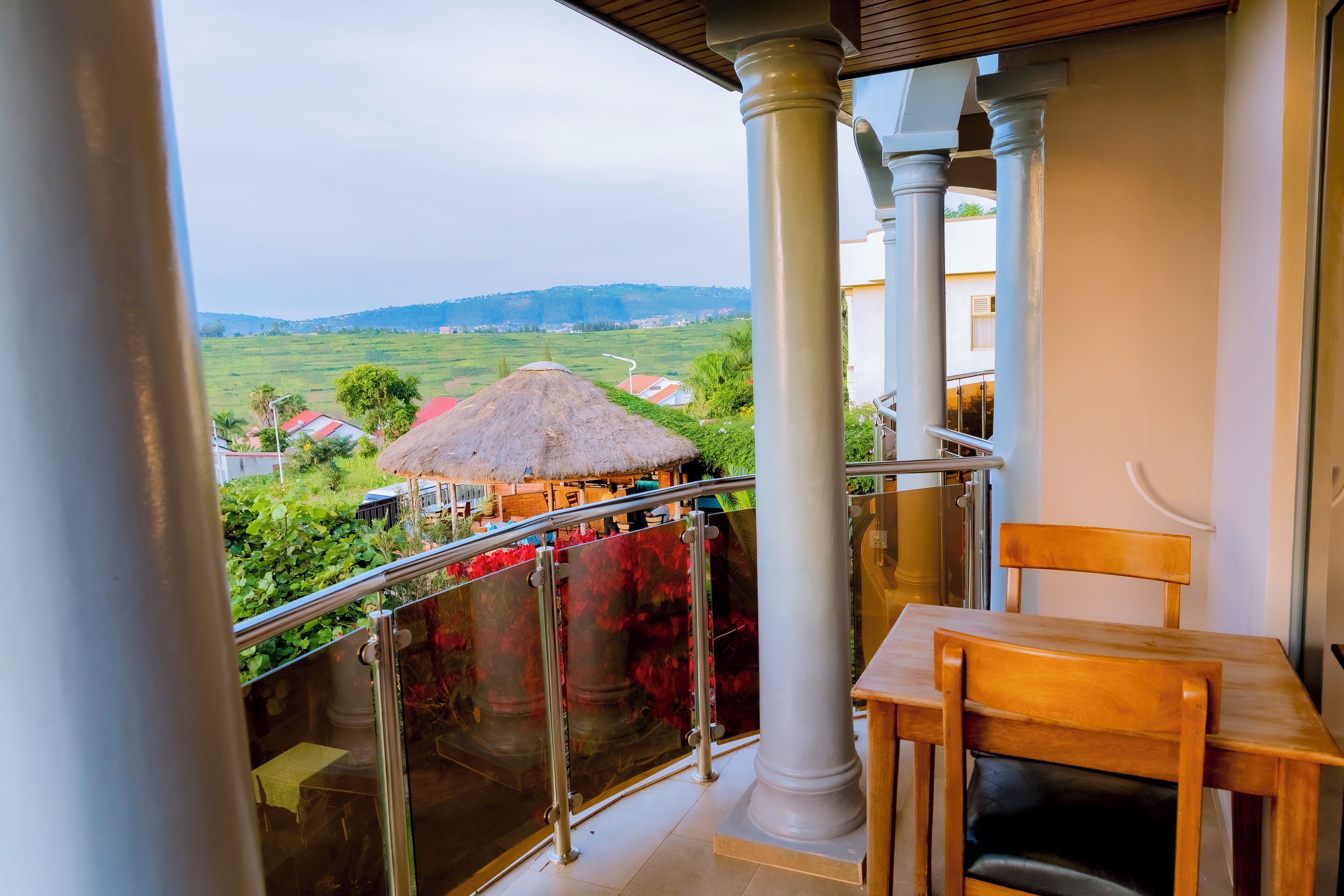 A balcony with a table and chair, a glass railing, and a view of a rural landscape with houses and a thatched-roof structure.