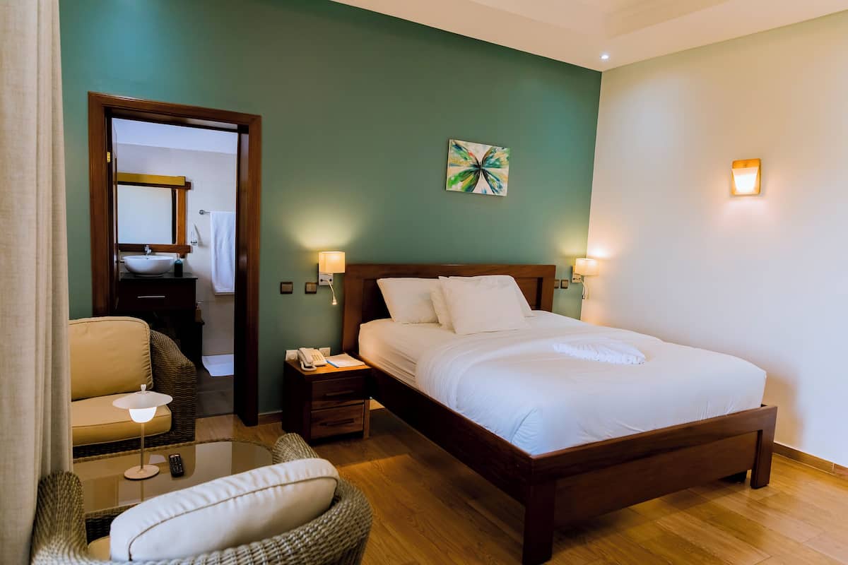 A hotel room with a bed, bedside tables, a chair, a small table, a mirror, and a painting on the wall.