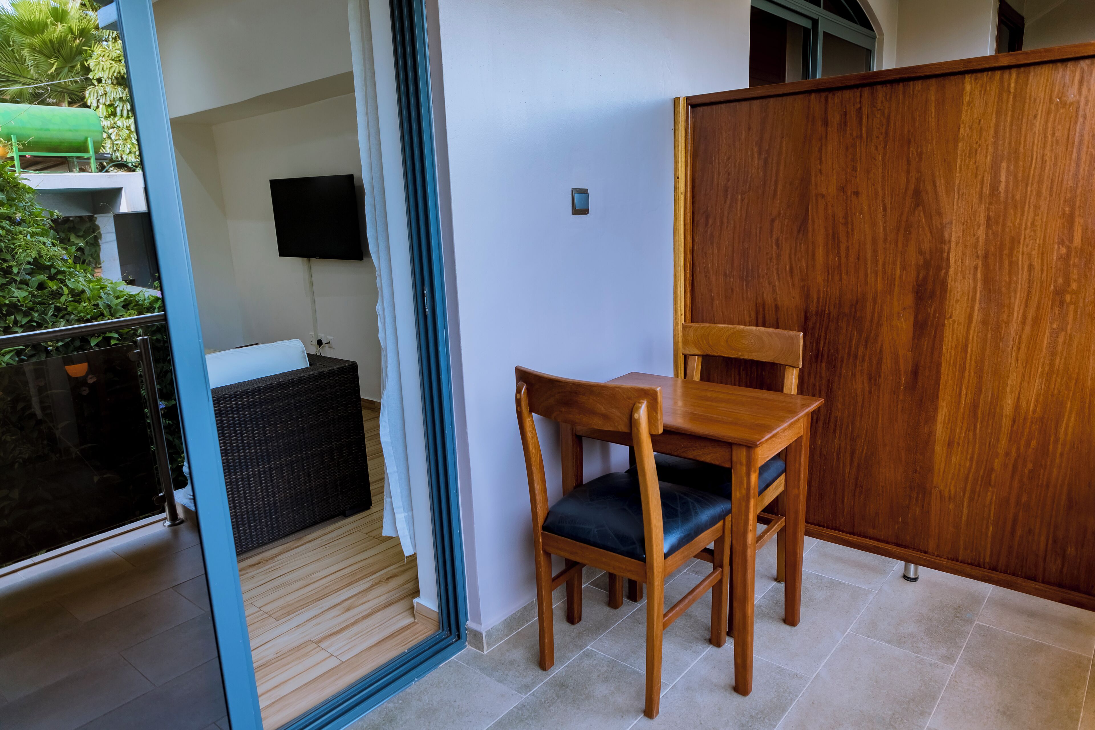 A room with a wooden dining table and chairs, a television mounted on the wall, and a sliding glass door leading to an outdoor area with plants.