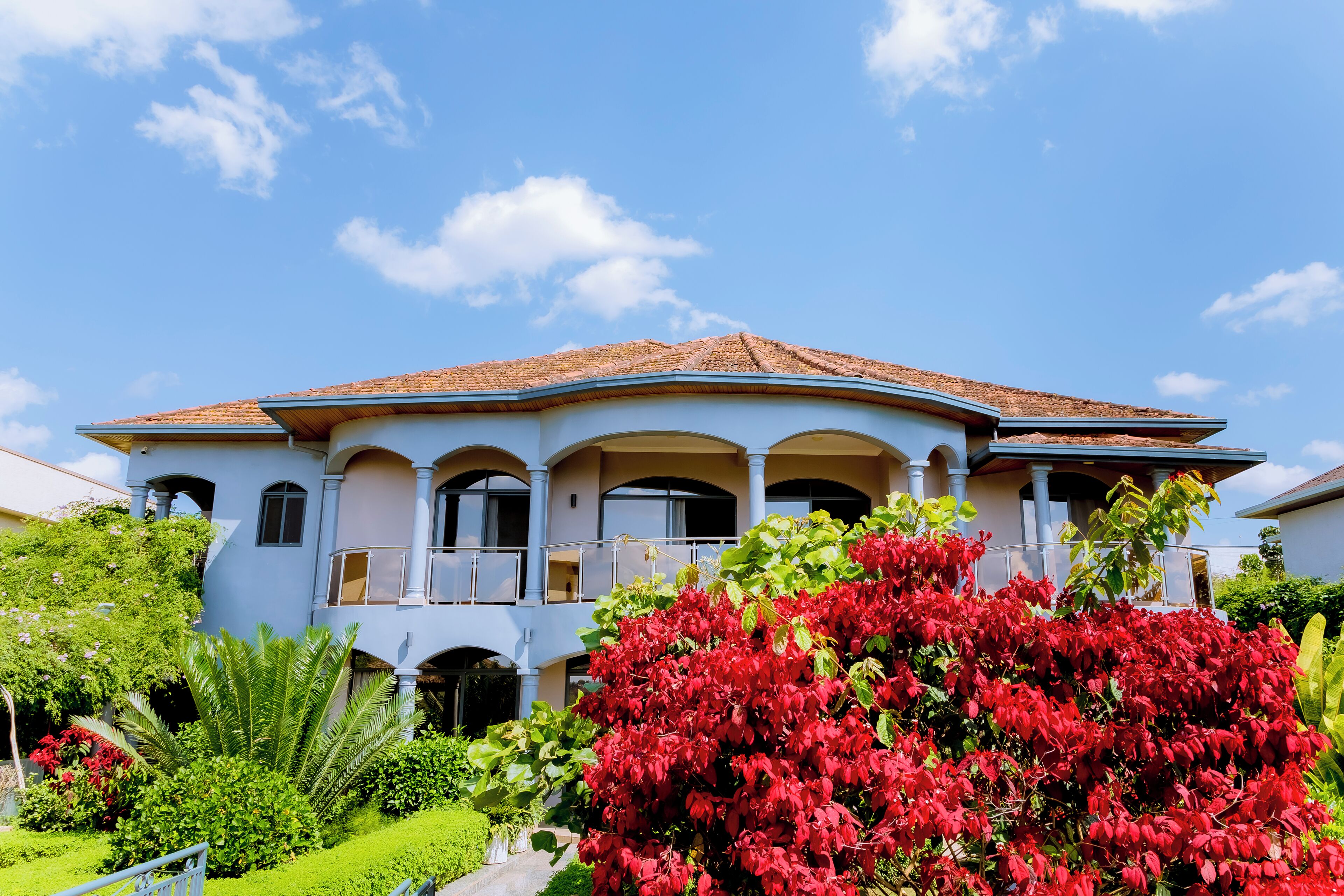 A two-story house with a tiled roof, multiple balconies, and a well-maintained garden with red flowering plants.