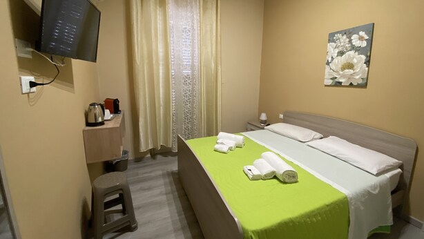 A hotel room with a bed, a television mounted on the wall, a desk with a kettle and a cup, a small stool, and a painting on the wall. - L'ERBA DEI SOVRANI (Palermo)