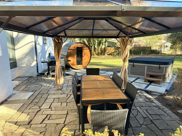 Una terrazza esterna con tavolo e sedie in legno, una zona relax coperta, una botte di legno e un camino in pietra.
