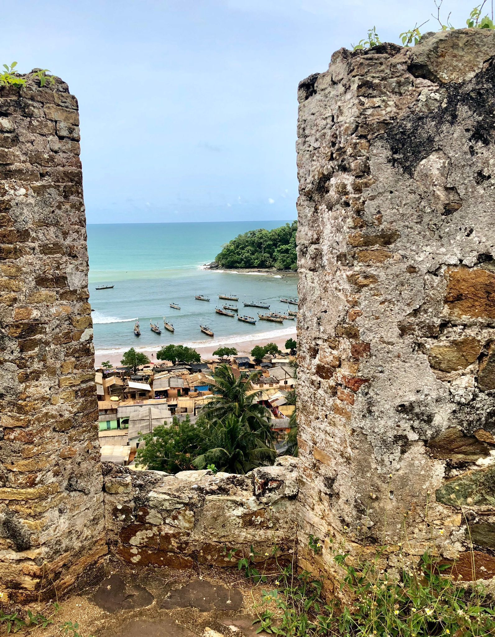 Vista através de antigas muralhas de pedra, com uma praia, barcos e uma pequena cidade.