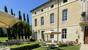 A well-maintained outdoor patio area with a wooden table and chairs, surrounded by neatly trimmed hedges and tall trees. - Chateau en Provence (Loriol-du-Comtat)