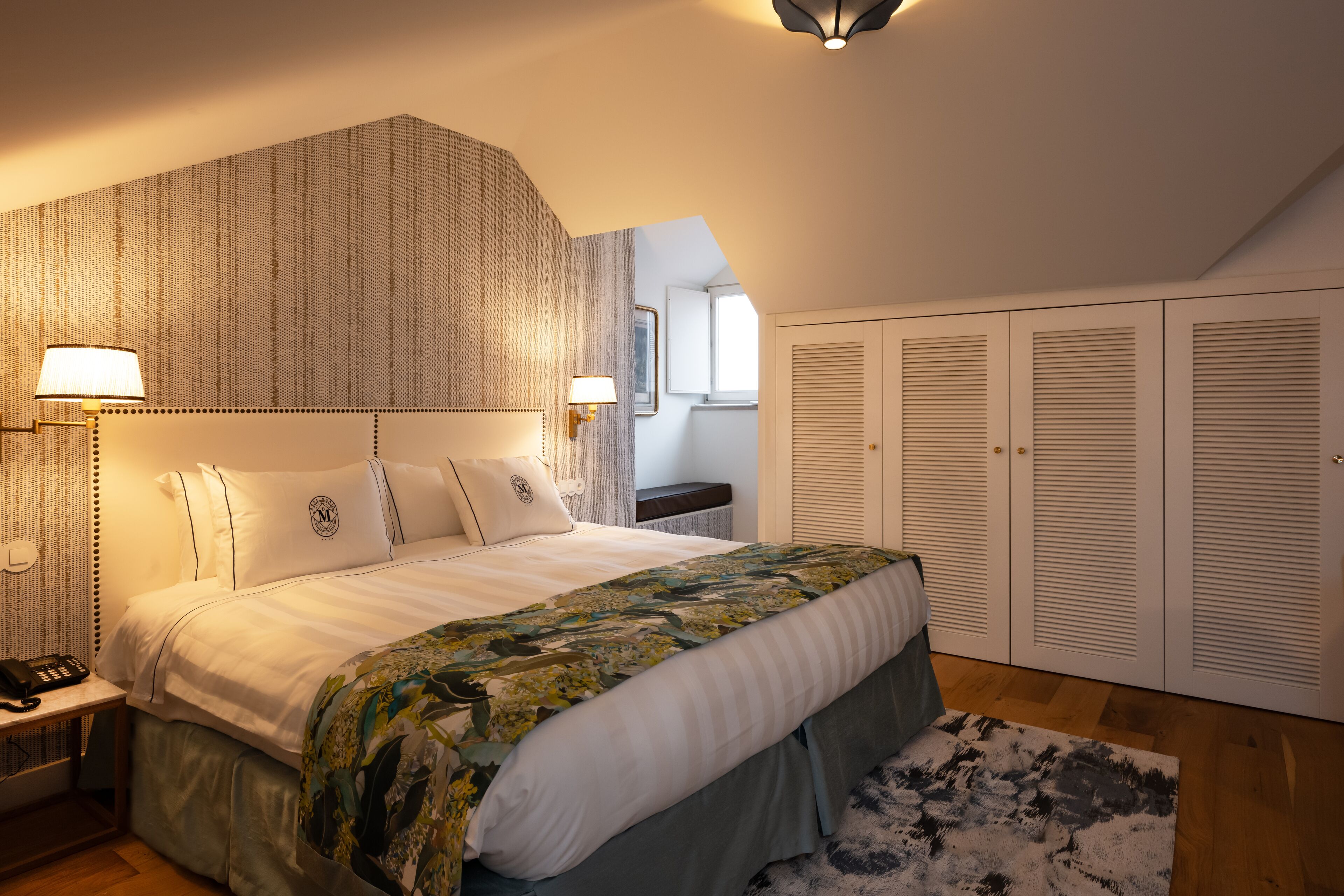 A bedroom with a large bed, a headboard with a textured pattern, bedside lamps, a bedside telephone, a window, and a set of closets.