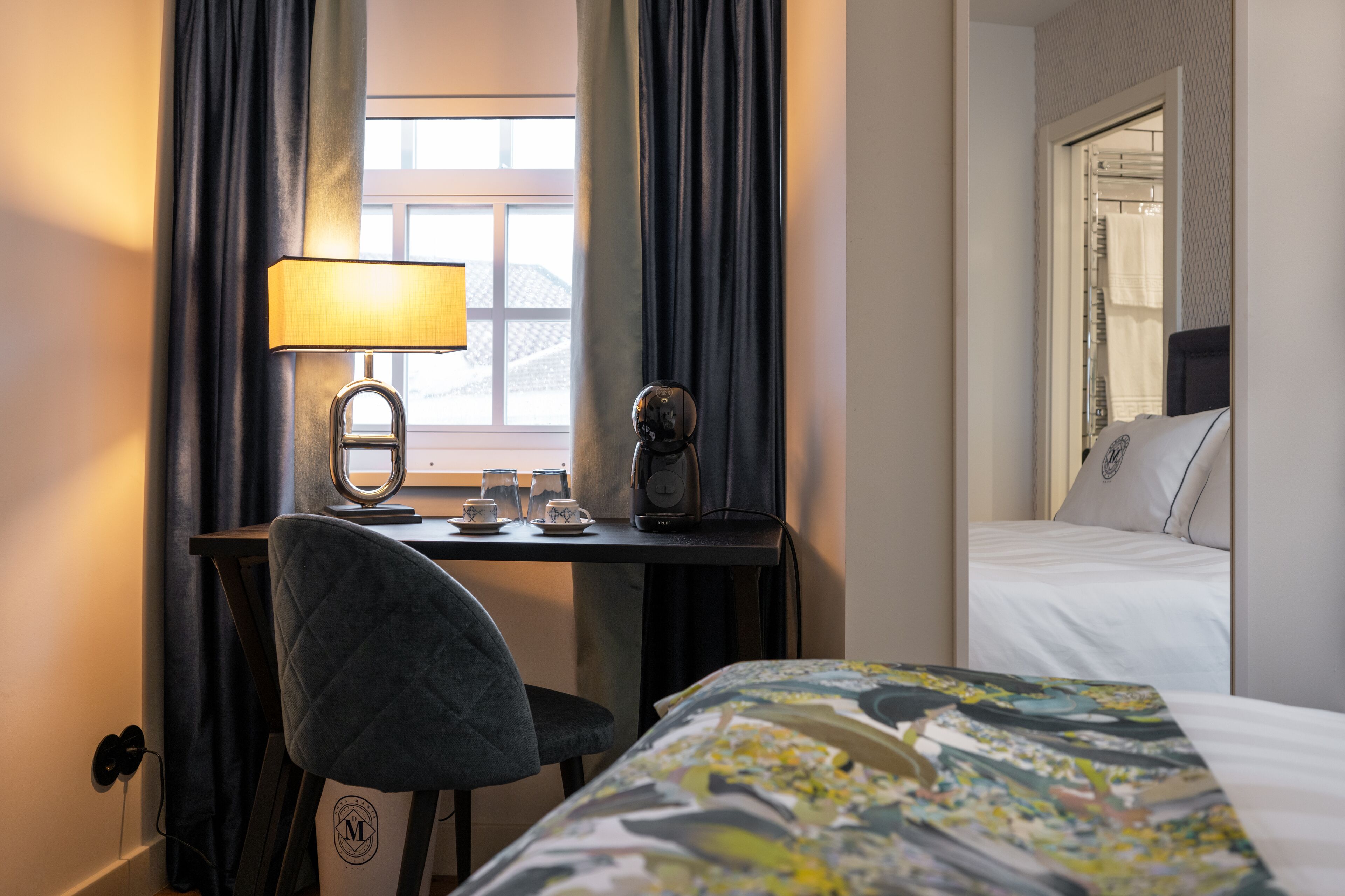 A hotel room with a bed, a desk with a lamp, a chair, and a window with curtains.