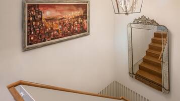 Un escalier avec une rampe en bois et un mur blanc. Au-dessus de l’escalier, on aperçoit un tableau encadré représentant un paysage urbain, et à droite, un miroir avec un cadre décoratif.