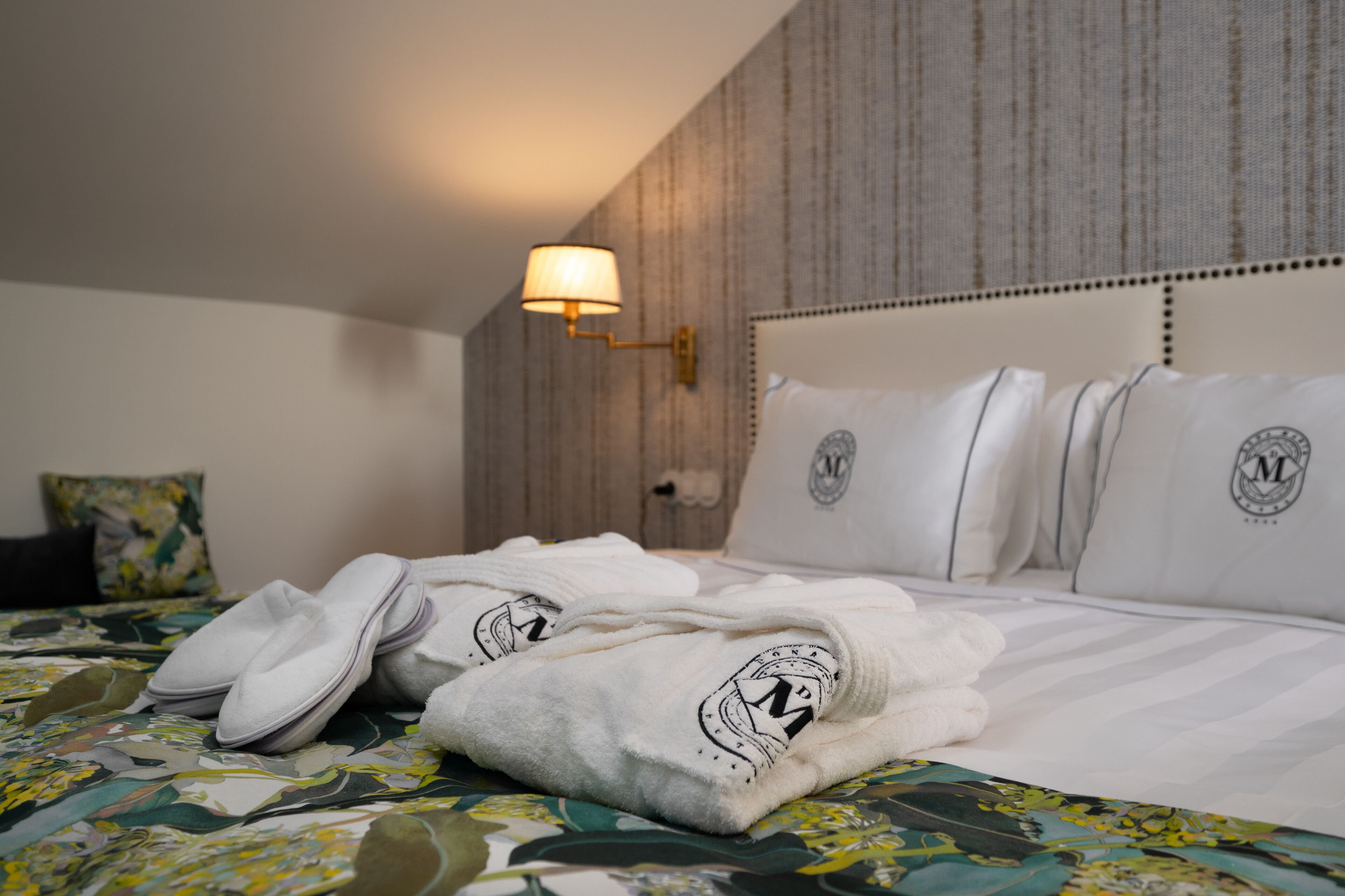 A neatly made bed with a floral bedspread, white pillows with a logo, and a pair of slippers on the bed.