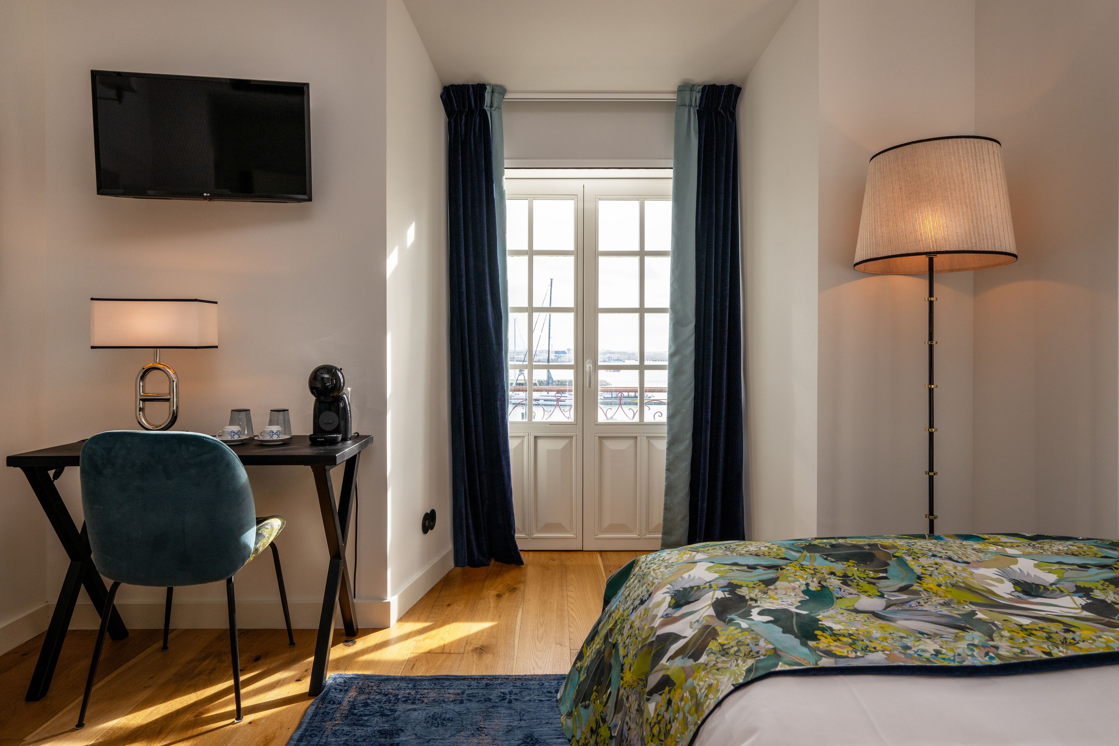 A bedroom with a bed, a desk with a chair, a TV mounted on the wall, and a window with curtains.
