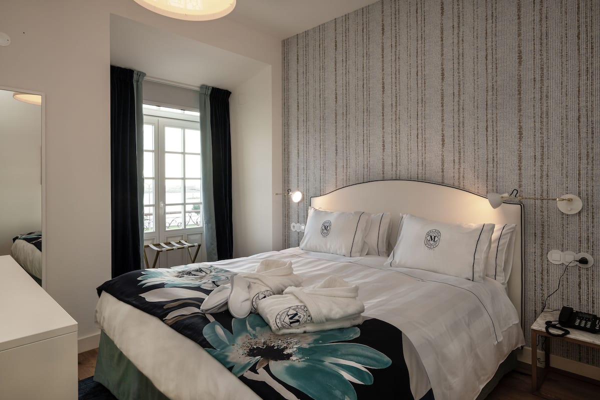 Ein ordentlich bezogenes Bett mit floralem Überwurf, ein Nachttisch mit Lampe und Blick ins Freie durch ein Fenster mit Vorhängen.