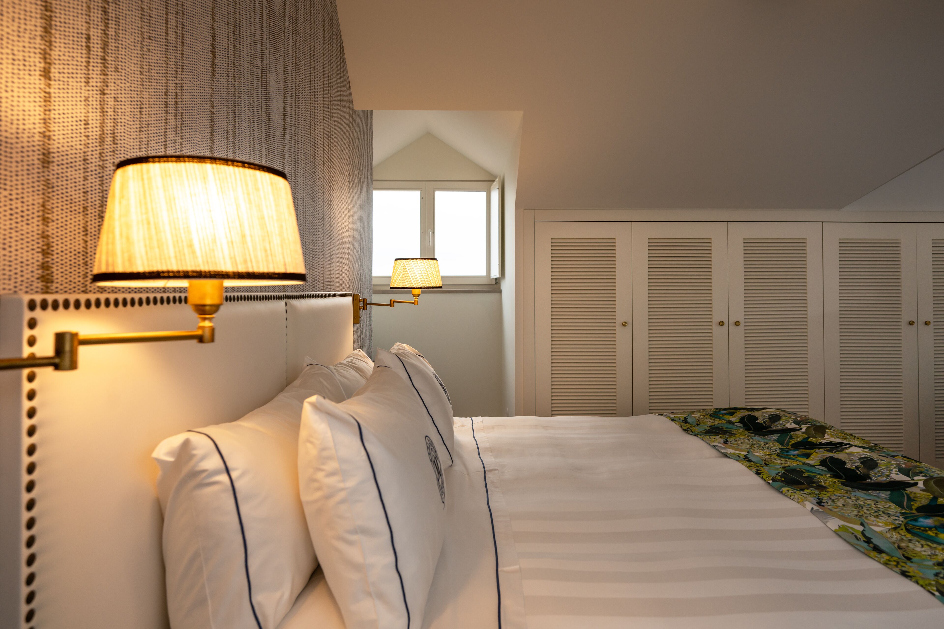 A neatly made bed with white linens and a patterned bedspread, a lamp with a beige shade, and a window with blinds.