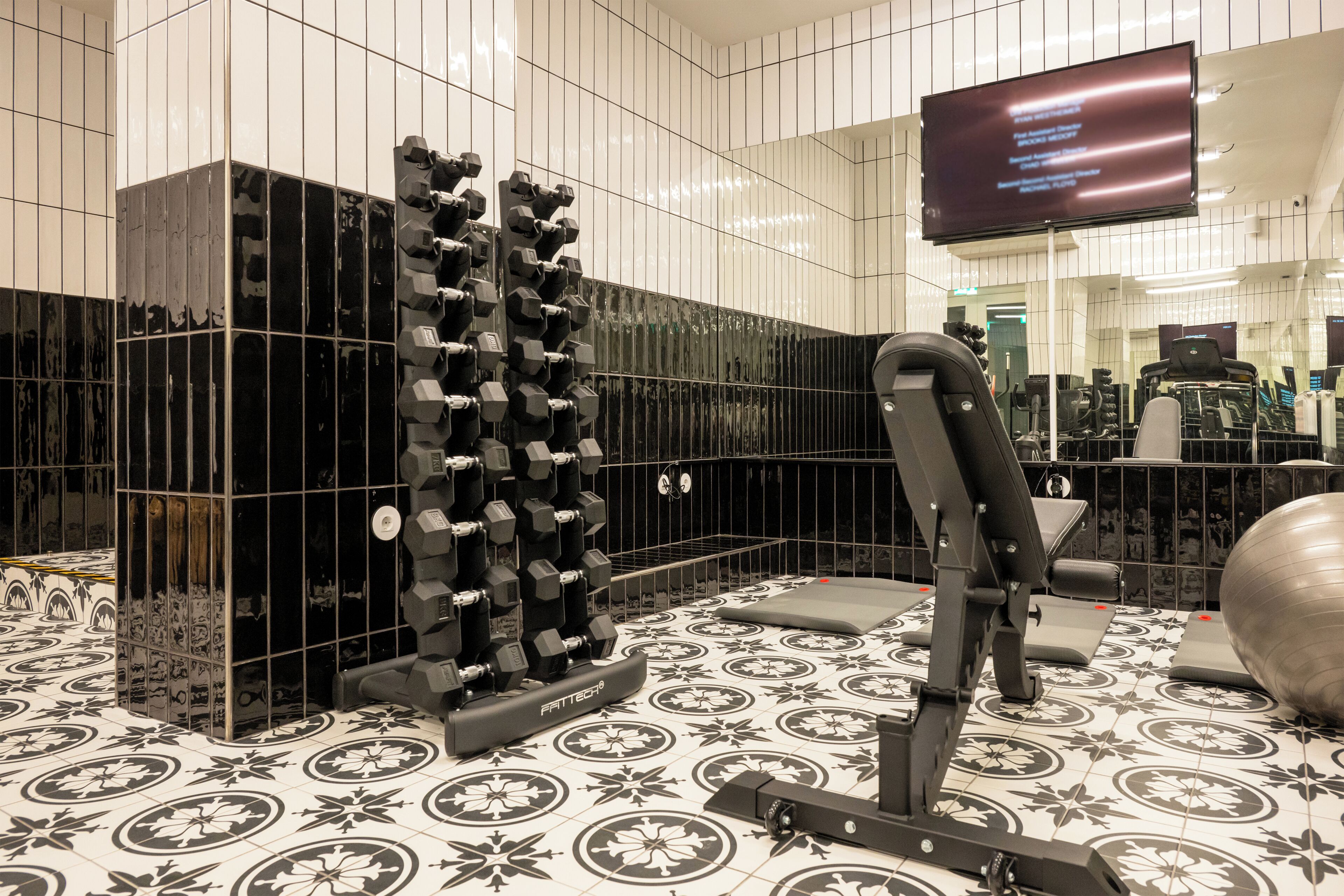 Une salle de sport équipée de matériel d’exercice, dotée d’un mur avec des miroirs et d’un téléviseur affichant une liste des équipements et services disponibles.